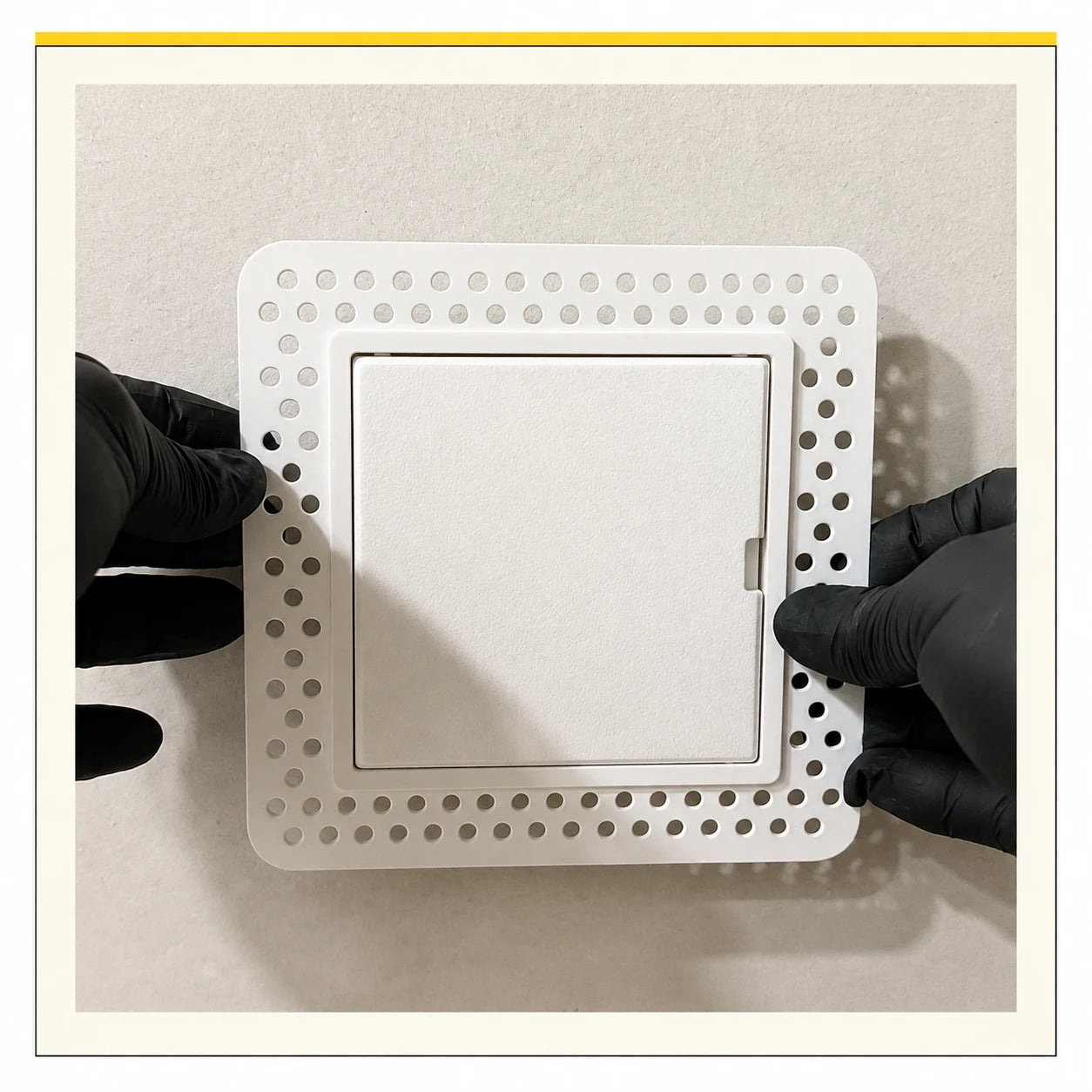 White square access panel with a perforated mud-in flange held against a wall by black-gloved hands