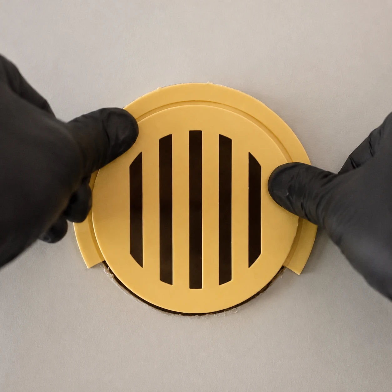 Yellow slotted round vent grille being fitted into a wall by black-gloved hands